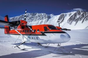 A red and white airplane with black stripes is parked on snow in a mountainous, icy landscape under a clear blue sky. Snow-covered peaks rise in the background—a scene typical of Antarctica and Antarctic Logistics and Expeditions.