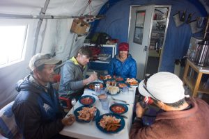 Four people sit around a table inside a tent at the South Pole, sharing a meal with plates of food and bowls of soup. The group appears relaxed and happy, dressed warmly amidst their Antarctic Logistics and Expeditions gear.
