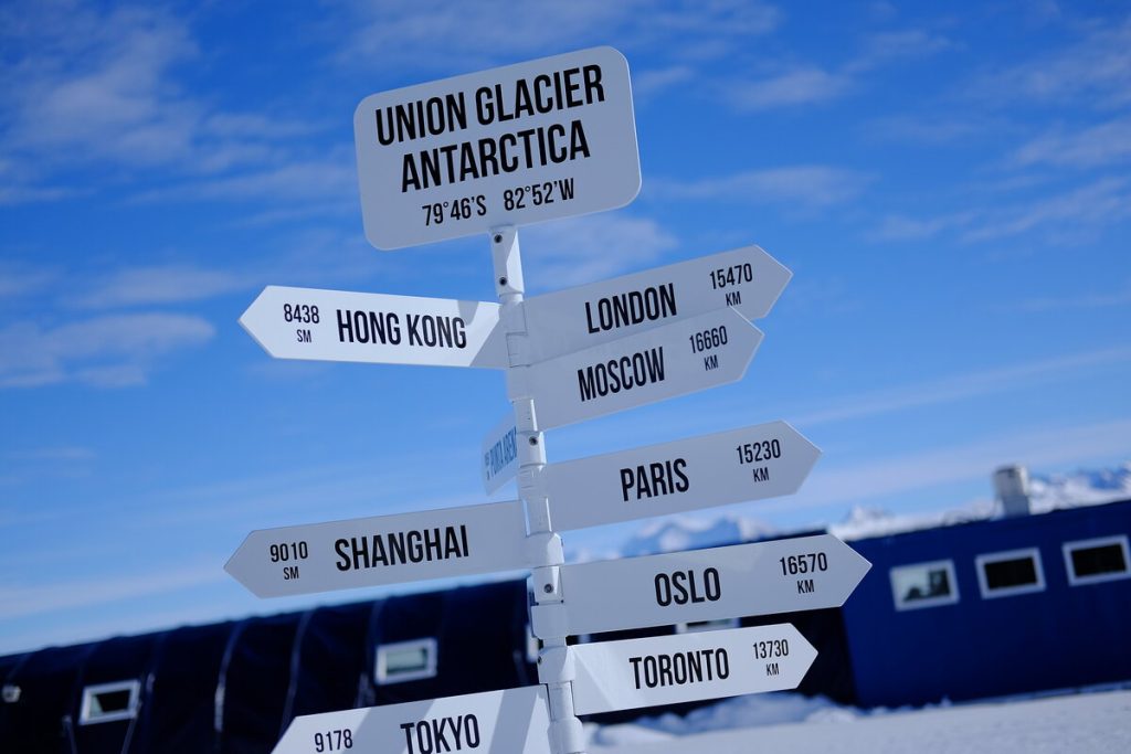 A signpost at Union Glacier, Antarctica, set up by Antarctic Logistics and Expeditions, shows distances to cities like Hong Kong, London, and Tokyo against a snowy landscape near the South Pole under a clear blue sky.