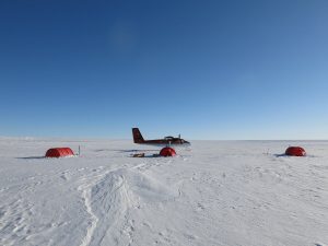 A small red aircraft is parked on a vast, snowy landscape in Antarctica under a clear blue sky, with three red tents set up nearby—typical of Antarctic Logistics and Expeditions operations.