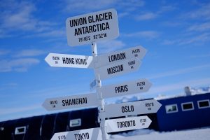A white signpost at Union Glacier, Antarctica, managed by Antarctic Logistics and Expeditions, shows distances to cities like Hong Kong, London, and Toronto, with snowy mountains and blue sky near the South Pole in the background.