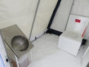 A metal sink with a water container underneath is on the left, and a white portable toilet with an open lid is on the right, both set up inside a tent on a snowy surface at the South Pole with Antarctic Logistics and Expeditions.