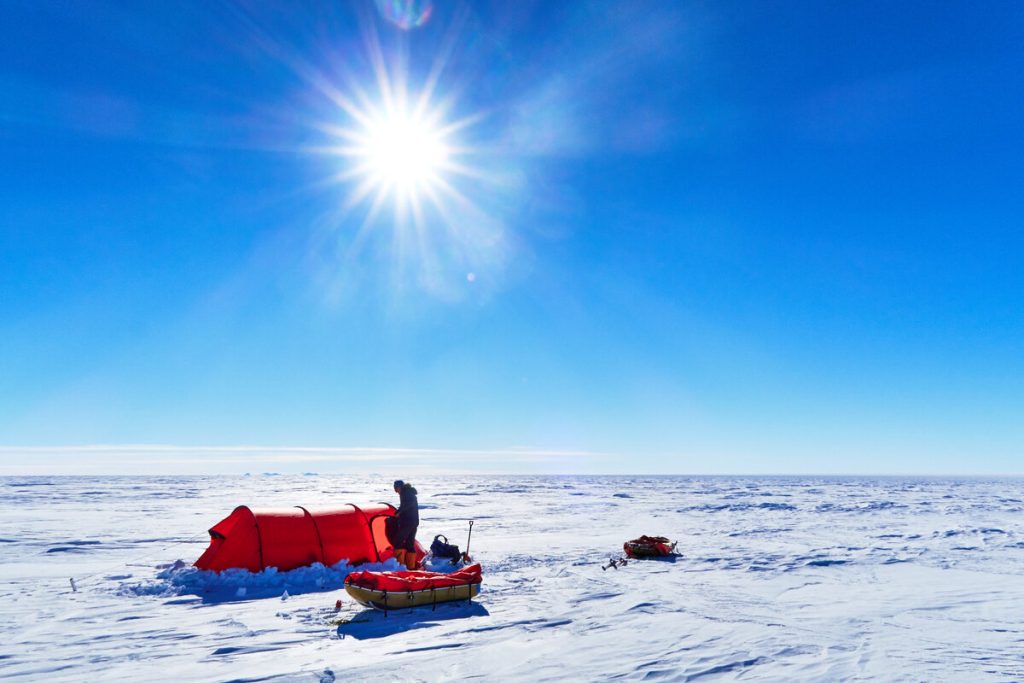 A person stands beside a red tent and supplies on the vast, snowy landscape of Antarctica under a bright sun and clear blue sky, with shadows cast on the snow—a scene reminiscent of Antarctic Logistics and Expeditions at the South Pole.