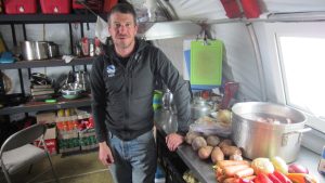 A man stands in a kitchen tent at the South Pole next to a counter with potatoes, carrots, onions, and a large pot. Shelves behind him hold kitchen supplies, soda bottles, and food items as sunlight streams through a window on the right.