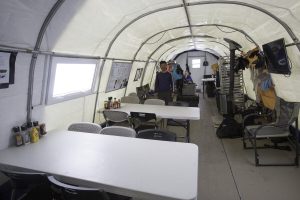 A large tent interior with folding tables and chairs lined up, typical of Antarctic Logistics and Expeditions at the South Pole. Two people stand at the back as shelves hold clothing and gear, while condiment bottles sit on a table in the foreground.