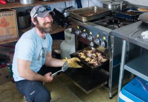 A smiling man wearing a blue shirt and cap uses an oven mitt to remove a tray of cooked meat from an oven in a busy kitchen, reminiscent of life at Antarctic Logistics and Expeditions' South Pole base. Cooking utensils, pans, and boxes are visible in the background.