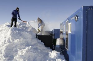 Two people wearing winter clothing shovel snow from a large mound beside blue portable buildings under a clear sky at the South Pole. Steam or snow particles drift in the cold Antarctic air.