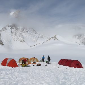 Several colorful tents are set up on a snowy mountain slope in Antarctica, with people in winter gear nearby and snow-covered peaks rising in the background—an adventure made possible by Antarctic Logistics and Expeditions.