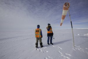 Two people in orange safety vests stand on snow at the South Pole near a wind sock, with cloudy skies above and the endless Antarctic landscape stretching into the distance—an impressive scene of Antarctic Logistics and Expeditions.
