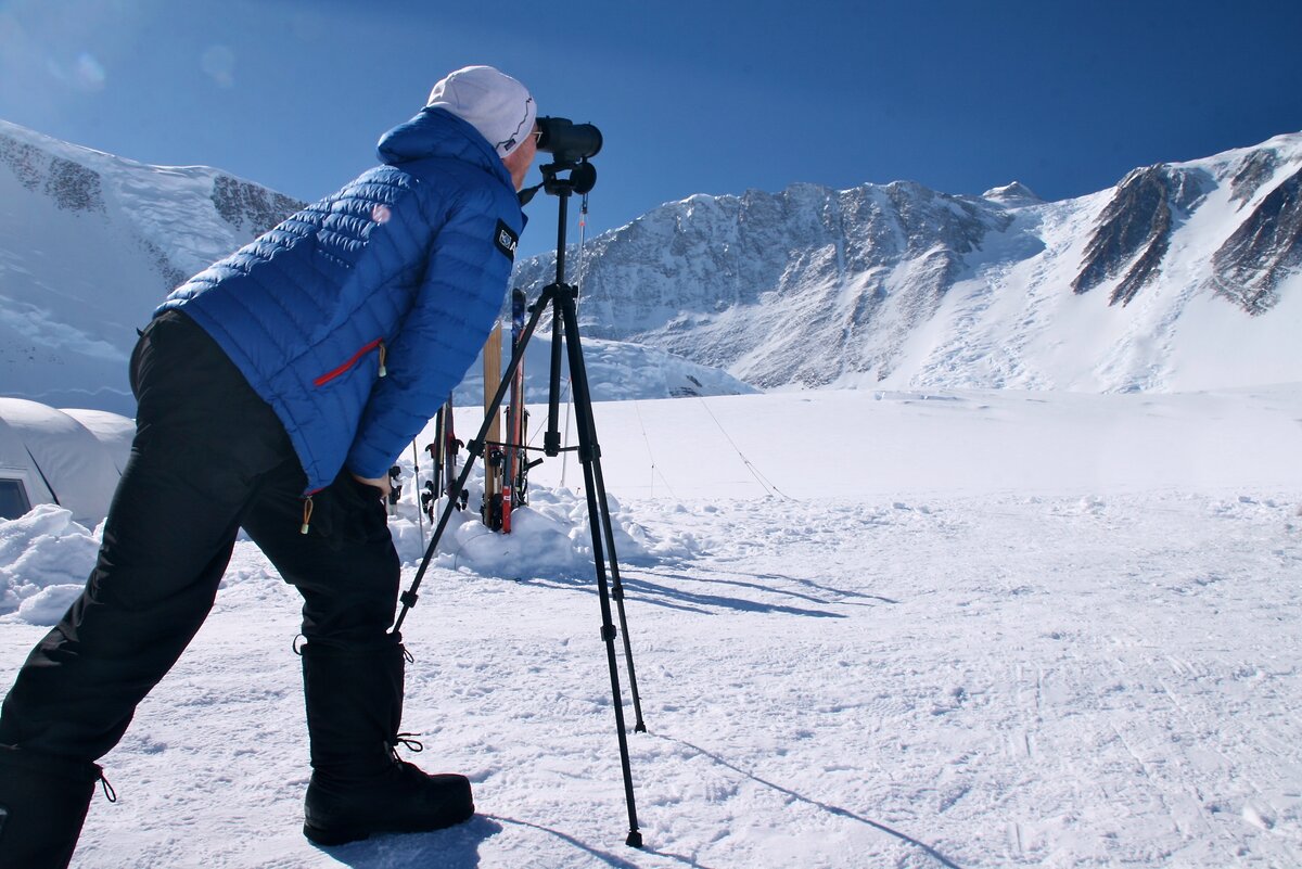 A person in winter gear looks through a spotting scope on a tripod, surveying the snowy, mountainous landscape of Antarctica under a clear blue sky.