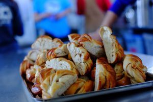 A tray filled with neatly arranged braided breads sits on a metal counter at an Antarctic Logistics and Expeditions kitchen, with blurred people and South Pole activity in the background.