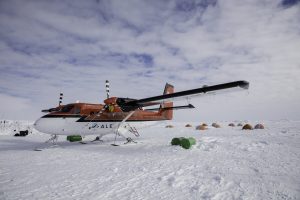 A red and white airplane with skis is parked on snowy ground near green barrels in Antarctica. Several orange tents, part of an Antarctic Logistics and Expeditions camp, are set up in the background under a partly cloudy sky.
