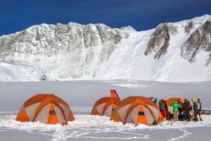 Several orange tents are set up on snow near a group of people in winter clothing, with the snow-covered mountains of Antarctica and a clear blue sky in the background—an expedition staged by Antarctic Logistics and Expeditions.