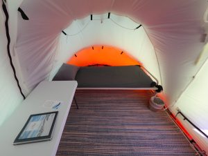 Interior of a tent with a single bed and pillow, striped rug, small white table with documents and pen, and trash bin. Soft orange light glows from the tent’s back wall at an Antarctic Logistics and Expeditions South Pole camp.