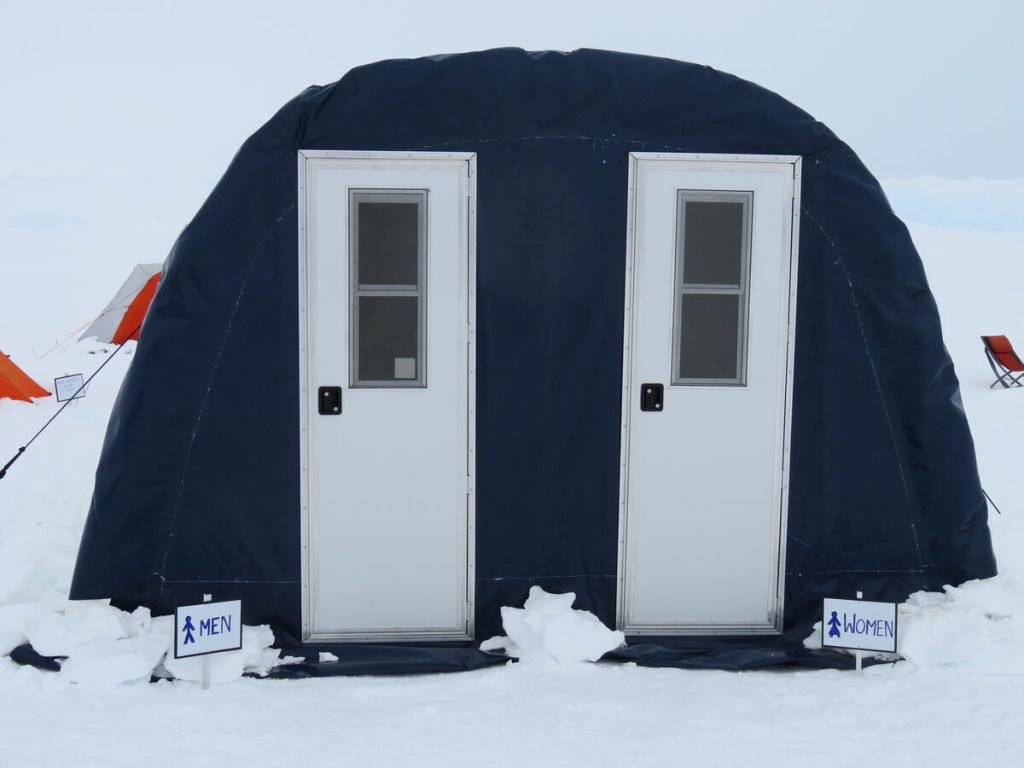A dark blue tent-like structure in snowy surroundings at the South Pole, with two white doors side by side. Signs in front indicate entrances for men and women. Orange tents and chairs are visible in the background.