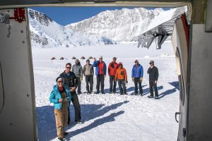 A group of people in winter gear stand on snowy ground near several tents, surrounded by snow-covered mountains in Antarctica, as seen from the open doorway of an Antarctic Logistics and Expeditions aircraft.
