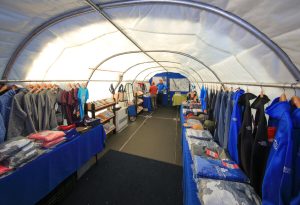 A man stands at the end of a long, white tent filled with tables and racks displaying shirts, jackets, hats, and South Pole souvenirs for sale. The merchandise is neatly arranged on both sides of the Antarctic Logistics and Expeditions tent.