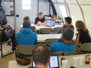 A group of people sit at tables inside a tent in Antarctica, attentively listening to a presenter from Antarctic Logistics and Expeditions, who points to a laptop screen displaying an image. Posters and equipment are visible around the tent.