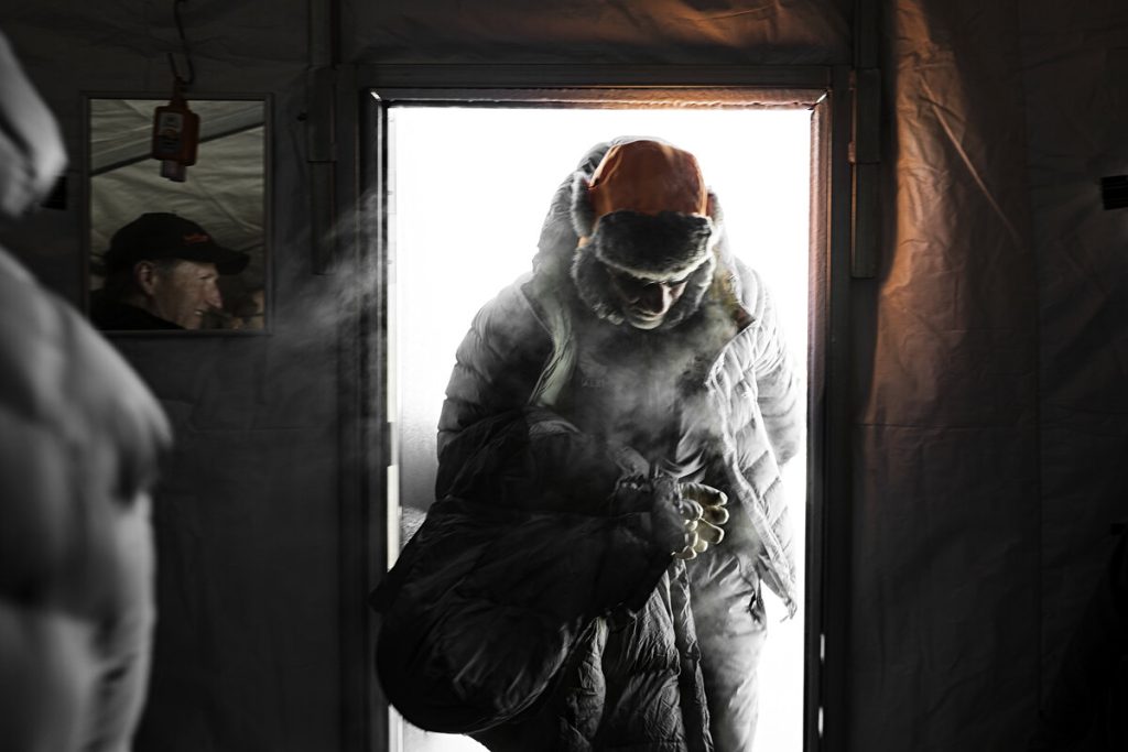 A person wearing a heavy winter coat and fur hat enters through a door at an Antarctic Logistics and Expeditions base, exhaling visible breath in the South Pole cold. Bright light from Antarctica contrasts with the dim, insulated indoor space.
