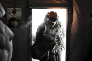 A person wearing a heavy winter coat and fur hat enters through a door at an Antarctic Logistics and Expeditions base, exhaling visible breath in the South Pole cold. Bright light from Antarctica contrasts with the dim, insulated indoor space.