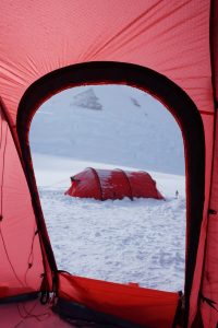 View from inside a red tent looking out at another red tent set up on a snowy Antarctic landscape with a mountain in the background. Snow blankets the ground, capturing the cold, wintry atmosphere typical near the South Pole.