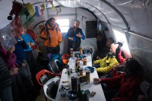 A group of people gather inside a tent around a long table covered with food, drinks, and supplies. Some are sitting, others standing in outdoor gear. Prayer flags hang above and light streams in—typical camaraderie on an Antarctic Logistics and Expeditions trip.