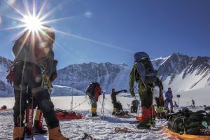 A group of mountaineers with backpacks and climbing gear stand on a snowy plateau in Antarctica, preparing for an Antarctic Logistics and Expeditions journey under bright sunlight, with rugged, snow-covered mountains in the background.