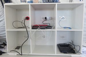 A white cubby shelf with six compartments, each containing electrical outlets, various charging cables, laptops, tablets, and a book titled World Atlas on top—perfect for planning journeys to Antarctica or South Pole expeditions.