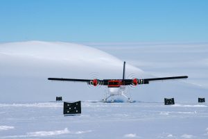 A small red-and-white propeller plane is parked on a snowy landscape in Antarctica, with white hills and a blue sky in the background; several black markers with white circles stand in the snow near the plane.