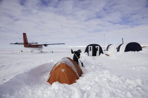 A person stands near an orange tent, surrounded by snow at an Antarctic Logistics and Expeditions camp, with a small red airplane and larger tents in the background under a partly cloudy sky, suggesting a remote South Pole expedition.