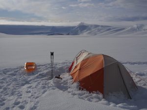 An orange and beige tent is set up on a snowy landscape in Antarctica with two ski poles and a small orange sled nearby. Snow-covered mountains and a cloudy sky, typical of Antarctic Logistics and Expeditions, are visible in the background.