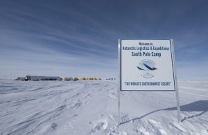A sign reads “Welcome to Antarctic Logistics and Expeditions South Pole Camp, The World’s Southernmost Resort.” In the snowy expanse of Antarctica, tents and structures stand beneath a wide, cloudy sky.