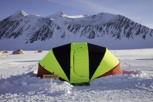A brightly colored yellow and black tent labeled Shackleton sits on snowy ground in Antarctica, with snow-covered mountains and more tents—part of Antarctic Logistics and Expeditions—visible in the background under a clear sky.
