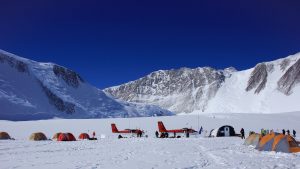 A snowy mountain camp scene in Antarctica with colorful tents and two red airplanes on the snow, set against rugged, snow-covered peaks under a clear blue sky. People with Antarctic Logistics and Expeditions gather near the tents and planes.