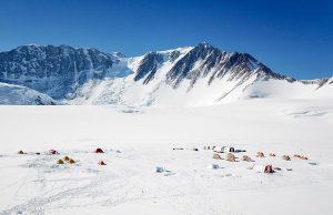 A snowy mountain range rises behind a field of tents set up on Antarctica’s vast, flat expanse of snow under a clear blue sky, suggesting a remote South Pole expedition or research camp.