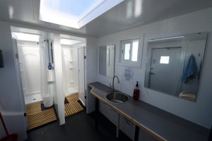 A modern, clean bathroom reminiscent of Antarctic Logistics and Expeditions features a shower stall, toilet, long countertop, sink, mirror, small window, and door. Towels hang by the door and wooden mats sit on the floor near the shower.
