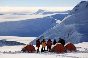 A group of people in heavy winter gear stand beside four orange tents on a snowy mountain slope, with vast snow-covered peaks and dramatic ridges in the background—a classic scene from an Antarctica expedition.