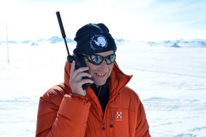 A person in an orange jacket and black beanie talks on a satellite phone while standing on a snowy, sunlit Antarctic landscape with mountains in the background, capturing the spirit of exploration in Antarctica.