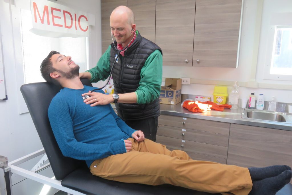 A patient in a blue sweater lies on an examination table, smiling, while a doctor listens to his chest with a stethoscope in a medical office—just another checkup before heading to the South Pole with Antarctic Logistics and Expeditions.