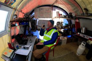 A person in a reflective vest and beanie sits inside a tent-like structure in Antarctica, smiling and speaking into a radio. The space, set up by Antarctic Logistics and Expeditions, is filled with cooking equipment, notes, and electronics.