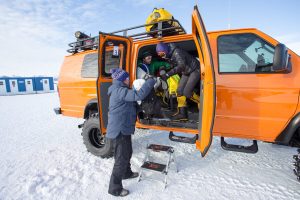 Three people in winter gear help each other get in and out of a bright orange Antarctic Logistics and Expeditions van parked on snowy ground, with portable toilets and snowy mountains in Antarctica visible in the background.