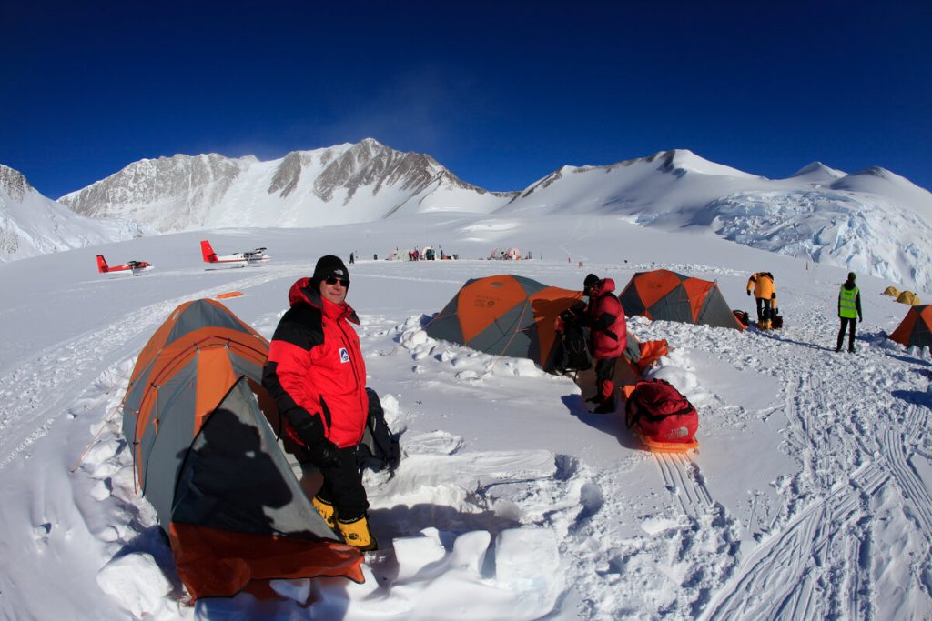 People in heavy winter gear set up bright orange tents on a snowy plateau surrounded by mountains in Antarctica; two small red planes from Antarctic Logistics and Expeditions are parked in the background under a clear blue sky.