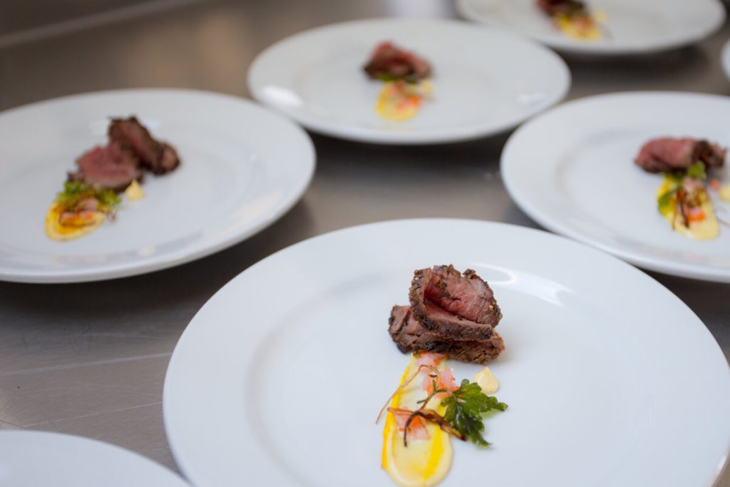 Several white plates with gourmet dishes—each featuring slices of seared beef, a swirl of yellow sauce, and fresh herbs—are arranged on a stainless steel countertop, showcasing fine dining even in the heart of Antarctica with Antarctic Logistics and Expeditions.
