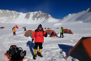 A person in a red jacket and yellow boots stands smiling and giving a thumbs up at an Antarctic Logistics and Expeditions campsite, with orange tents, snow-covered mountains, and a red aircraft set against the frozen expanse of Antarctica.