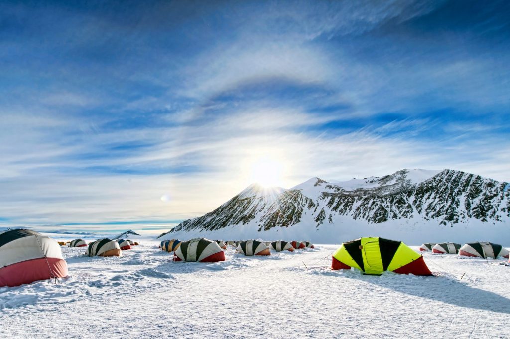 A camp with colorful tents is set up on a snowy landscape under a bright sky, with the sun rising or setting behind snow-covered mountains in Antarctica, capturing the spirit of Antarctic Logistics and Expeditions near the South Pole.