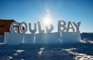 Large snow blocks spell “GOULD BAY” against a bright blue sky in Antarctica, with the sun shining behind the letters and casting long shadows. A tent is visible on the left—a striking scene supported by Antarctic Logistics and Expeditions.