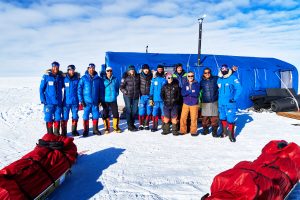 A group of people in cold-weather gear stand in front of a blue tent on snow in Antarctica, with supplies packed in red bags nearby, under a partly cloudy sky—part of an Antarctic Logistics and Expeditions journey.