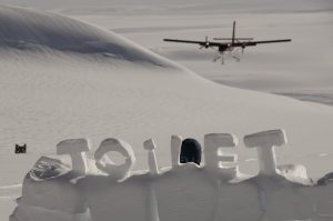 A red and white plane lands on a snowy landscape in Antarctica. In the foreground, large snow blocks spell out “TOILET,” with a blue toilet seat visible, showcasing the unique setups at Antarctic Logistics and Expeditions near the South Pole.