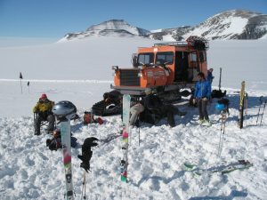 Several people in winter gear rest on snow beside an orange tracked vehicle from Antarctic Logistics and Expeditions, surrounded by skis and poles, with snowy mountains and blue sky hinting at the remote beauty of Antarctica.