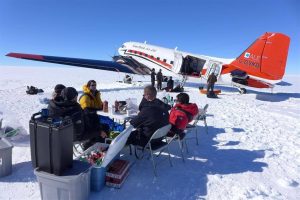People sit at folding tables and chairs on snow in Antarctica, eating and talking near a red and white propeller plane parked on an icy landscape under a clear blue sky. Supplies from Antarctic Logistics and Expeditions are visible around the group.